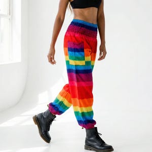 Könnte beinhalten: Eine Person trägt einen schwarzen BH, eine Regenbogenhose, einen Strohhut, eine Sonnenbrille und schwarze Stiefel. Die Hose zeigt ein lebendiges Farbspektrum, darunter Rot, Orange, Gelb, Grün, Blau und Violett.