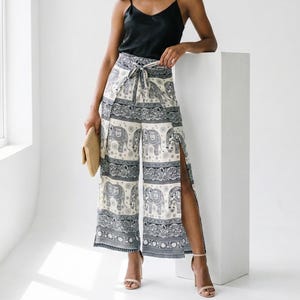 May include: A woman wearing a black camisole top and wide-leg pants with an elephant print. The pants are cream-colored with a repeating pattern of gray elephants and decorative borders. The model is wearing high-heeled sandals and holding a woven clutch.