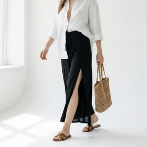 Puede incluir: Una mujer con camisa blanca, pantalones negros con abertura y sombrero de paja. Lleva un bolso tote tejido y sandalias marrones. El texto "Home 64 Studio" y "MODEL TALL 175 CM / 5.9 FEET" son visibles.