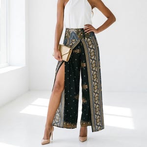 Könnte beinhalten: Eine Frau trägt ein weißes Neckholder-Top und eine gemusterte Palazzo-Hose mit hohem Schlitz. Sie hält eine goldene Clutch und trägt nudefarbene Absätze. Die Hose hat einen schwarzen Hintergrund mit einem detaillierten Muster. Der Text "Home 64 Studio" und "MODEL TALL 175 CM / 5.9 FEET" sind sichtbar.