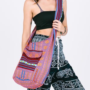 May include: A colorful shoulder bag with a patterned design in shades of pink, blue, and red. The bag has a long strap and a pocket on the front. The person is wearing black pants with an elephant print.