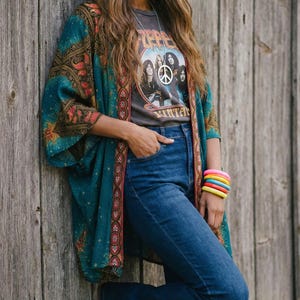May include: A teal kimono with a paisley design is worn over a gray graphic tee and blue jeans. The tee features a band image and a peace sign necklace. A stack of colorful bracelets and yellow sunglasses complete the look.