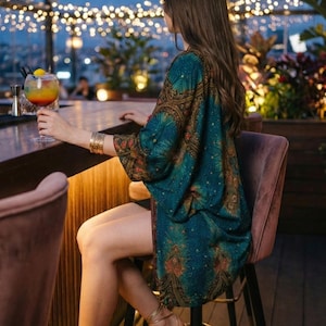 May include: A person seated at a bar, wearing a teal and brown patterned kimono, gold bracelets, and strappy heels. A colorful cocktail is held in a glass. The scene is set outdoors at night, with string lights in the background.