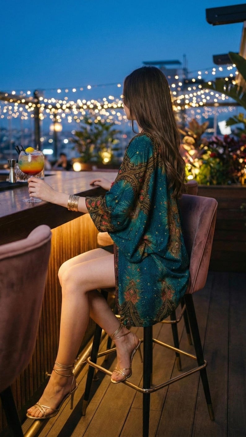 May include: A person seated at a bar, wearing a teal and brown patterned kimono, gold bracelets, and strappy heels. A colorful cocktail is held in a glass. The scene is set outdoors at night, with string lights in the background.