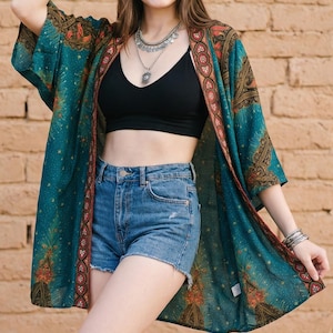 May include: A teal kimono with a paisley pattern and red trim is worn over a black top and denim shorts. The outfit is accessorized with a brown hat, sunglasses, and silver bracelets. Brown cowboy boots complete the look.