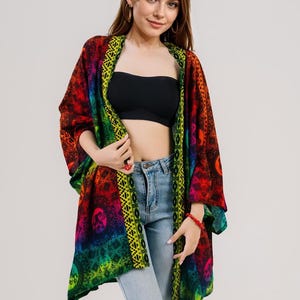 Pu&ograve; includere: Kimono aperto dai colori vivaci con un motivo arcobaleno. Il capo presenta un mix di tonalit&agrave; rosse, arancioni, gialle, verdi e blu, con un bordo decorativo. Indossato sopra un top nero e jeans azzurri.