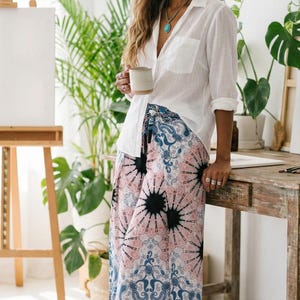 Chic indoor styling of the Pink Feather Palazzo Pants. A tall beautiful model stands by a rustic wooden table and green house plants, holding a cozy mug. She pairs these flowy boho summer beach vacation wear trousers with a classic white button-down shirt, a turquoise necklace, and beige mules. The vibrant pink print and deep blue paisley hem create a perfect relaxed yet sophisticated artistic everyday outfit! ☕🌿🎨