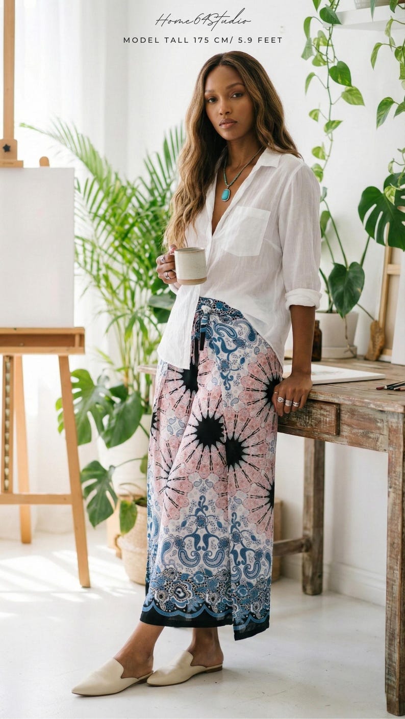 Chic indoor styling of the Pink Feather Palazzo Pants. A tall beautiful model stands by a rustic wooden table and green house plants, holding a cozy mug. She pairs these flowy boho summer beach vacation wear trousers with a classic white button-down shirt, a turquoise necklace, and beige mules. The vibrant pink print and deep blue paisley hem create a perfect relaxed yet sophisticated artistic everyday outfit! ☕🌿🎨
