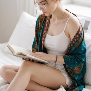 May include: A woman wearing a teal and orange patterned shawl, white tank top, and glasses reads a book while sitting on a bed. She is wearing a silver necklace and a blue bracelet. The bed has white sheets and pillows.