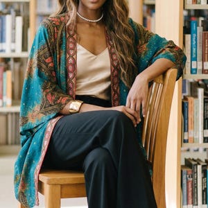 May include: A woman seated in a library setting, wearing a teal kimono with a floral pattern, a gold top, and black pants. She is also wearing a headband, pearl necklace, and gold bracelet. The background includes bookshelves.