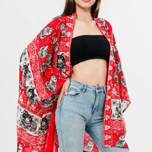 Pu&ograve; includere: Un kimono rosso con stampa di gatti in bianco e nero. Il capo &egrave; aperto sul davanti e ha maniche ampie. La modella indossa un top nero e jeans azzurri.