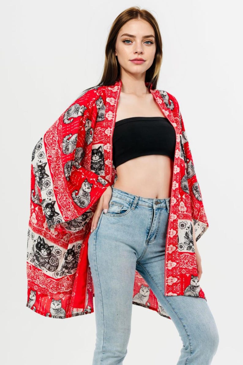 Pu&ograve; includere: Un kimono rosso con stampa di gatti in bianco e nero. Il capo &egrave; aperto sul davanti e ha maniche ampie. La modella indossa un top nero e jeans azzurri.