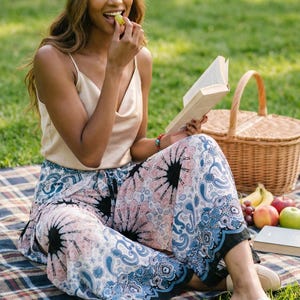 Cheerful tall model enjoying a sunny outdoor park picnic on a plaid blanket, wearing the gorgeous Pink Feather Palazzo Pants. These comfortable flowy boho summer beach vacation wear trousers are styled with a beige satin camisole, a cute cream beret, and slip-on loafers. She is smiling, eating fresh grapes, and reading a book next to a wicker picnic basket. The pink and blue paisley pants are perfect for relaxed weekend outings! 🍇🧺📚
