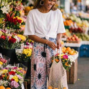 Happy tall woman shopping at a vibrant outdoor flower and fruit market wearing the lovely Pink Feather Palazzo Pants. These flowy boho summer beach vacation wear trousers are styled casually with a knotted white t-shirt, brown slide sandals, and a net market bag filled with fresh oranges and daisies. The gorgeous pink and blue paisley wide-leg pants are incredibly comfortable for everyday casual summer city strolls! 🍊🌻🛍️