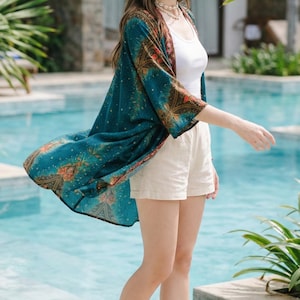 May include: A woman wearing a straw hat, sunglasses, and a teal kimono with a floral pattern. She is also wearing a white tank top, beige shorts, and sandals. The background features a swimming pool and tropical plants.