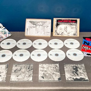 Puede incluir: Un conjunto de artículos de música punk rock, incluyendo CDs, parches y un folleto. Los CDs son blancos con un logo de calavera y huesos cruzados. Los parches son de varios colores con nombres de bandas. El folleto está abierto, mostrando ilustraciones.