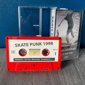 K&ouml;nnte beinhalten: Ein rotes Kassettenband mit dem Titel "Skate Punk 1998" und dem Text "Nomadic Pulse Records Presents..." Das Kassettenband befindet sich in einem durchsichtigen Plastikgeh&auml;use mit einem kleineren Geh&auml;use, das eine Liste von Songs enth&auml;lt.