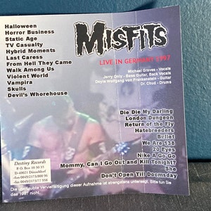 May include: A black and white Misfits concert poster with a photo of the band members. The poster lists the band's name, the concert date, the location, and the band members' names. The poster also lists the band's song titles, including "Die Die My Darling", "London Dungeon", "Return of the Fly", "Hatebreeders", "Bullet", "We Are 138", "20 Eyes", "Nike A Go Go", "Mommy, Can I Go Out and Kill Tonight?", and "She". The poster also includes the text "Don't Open Till Doomsday".