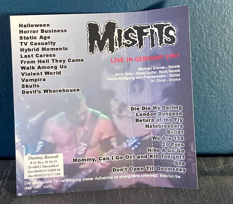 May include: A black and white Misfits concert poster with a photo of the band members. The poster lists the band's name, the concert date, the location, and the band members' names. The poster also lists the band's song titles, including "Die Die My Darling", "London Dungeon", "Return of the Fly", "Hatebreeders", "Bullet", "We Are 138", "20 Eyes", "Nike A Go Go", "Mommy, Can I Go Out and Kill Tonight?", and "She". The poster also includes the text "Don't Open Till Doomsday".