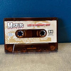 May include: A brown cassette tape with a gold label. The label has the text "Misfits Live in Germany 1997" and a list of songs. The cassette tape has a Dolby logo.