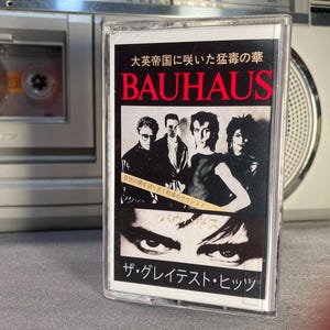 May include: A cassette tape with a black and white cover featuring the band Bauhaus. The cover has the band's name in red, Japanese text, and images of the band members. The cassette is in a clear plastic case.