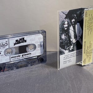 K&ouml;nnte beinhalten: Eine transparente Kassette des Albums "Music Giants" von Black Sabbath. Das Kassettengeh&auml;use ist transparent, mit dem Albumtitel und dem Bandnamen auf dem Etikett. Das Albumcover ist im Inneren sichtbar.