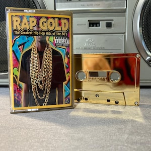May include: A gold cassette tape labeled "RAP GOLD: The Greatest Hip Hop Hits of the 80's" with a colorful album cover featuring a person wearing gold chains. A second gold cassette tape is next to it. A silver boombox is in the background.