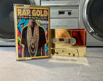 May include: A gold cassette tape labeled "RAP GOLD: The Greatest Hip Hop Hits of the 80's" with a colorful album cover featuring a person wearing gold chains. A second gold cassette tape is next to it. A silver boombox is in the background.