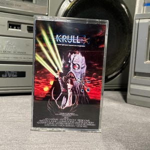 May include: A vintage cassette tape for the movie "Krull" with a colorful cover featuring a monster and a woman. The tape is in a clear plastic case, with the title and credits visible. The tape is set against a backdrop of vintage stereo equipment.