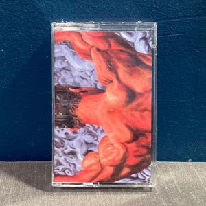 Könnte beinhalten: Eine Kassette mit einem roten und weißen Bild einer muskulösen Figur auf der Vorderseite. Die Figur ist mit erhobenen Armen über dem Kopf dargestellt.