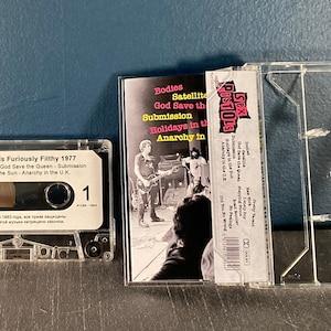 May include: A clear plastic cassette tape case with a white cassette tape inside. The cassette tape has a white label with the text "Sex Pistols Furiously Filthy 1977" and a list of songs: "Bodies", "Satellite", "God Save the Queen", "Submission", "Holidays in the Sun", and "Anarchy in the UK". The case has a picture of the band on the front with the text "Sex Pistols" in pink and black.