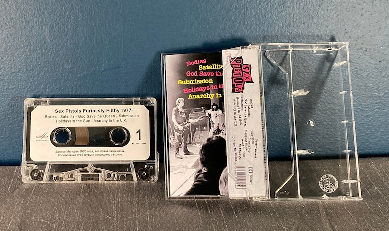 May include: A clear plastic cassette tape case with a white cassette tape inside. The cassette tape has a white label with the text "Sex Pistols Furiously Filthy 1977" and a list of songs: "Bodies", "Satellite", "God Save the Queen", "Submission", "Holidays in the Sun", and "Anarchy in the UK". The case has a picture of the band on the front with the text "Sex Pistols" in pink and black.