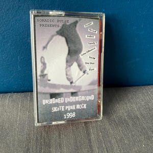 May include: A clear plastic cassette tape case with a black and white photo of a skateboarder. The text on the case reads "NOMADIC PULSE PRESENTS... UNSIGNED UNDERGROUND SKATE PUNK ROCK 1998".