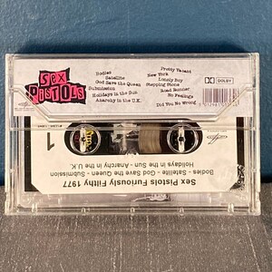 May include: A clear plastic cassette tape case with a black and white label. The label features the band name "Sex Pistols" in pink letters and a list of songs, including "God Save the Queen", "Anarchy in the U.K.", and "Holidays in the Sun". The cassette tape is labeled "Furiously Filthy 1977".