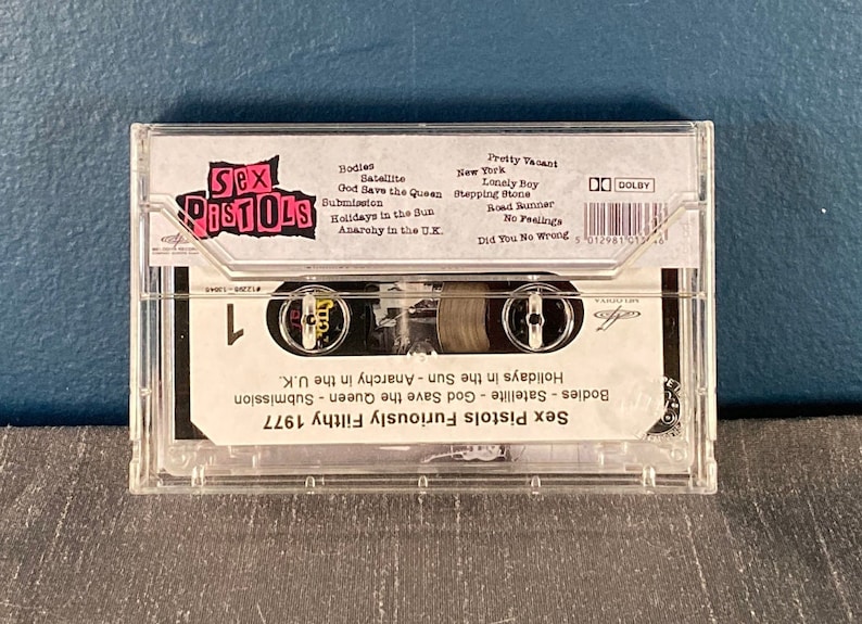 May include: A clear plastic cassette tape case with a black and white label. The label features the band name "Sex Pistols" in pink letters and a list of songs, including "God Save the Queen", "Anarchy in the U.K.", and "Holidays in the Sun". The cassette tape is labeled "Furiously Filthy 1977".