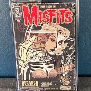 May include: A vintage-style concert poster for the Misfits, featuring a skull and a woman with a glass of liquid. The poster is in a clear plastic case. The text on the poster reads "Tales from the Misfits", "Durango", "Jughead", "11 Febrero", "Live!" and "26/29".