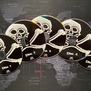Może przedstawiać: Pięć srebrnych płyt CD z czarno-białymi wzorami czaszek i skrzyżowanych kości. Płyty CD są oznaczone jako "Dysk 1", "Dysk 2", "Dysk 3", "Dysk 4" i "Dysk 5".