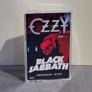 K&ouml;nnte beinhalten: Eine Musikkassette mit dem Album "Greatest Hits" von Black Sabbath. Das Cover zeigt den Namen "OZZY" in Wei&szlig;, eine rote Totenkopf-Grafik und den Bandnamen "BLACK SABBATH" in Wei&szlig;. Die Kassette befindet sich in einem transparenten Kunststoffgeh&auml;use.