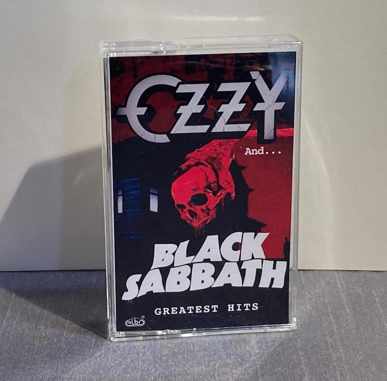 K&ouml;nnte beinhalten: Eine Musikkassette mit dem Album "Greatest Hits" von Black Sabbath. Das Cover zeigt den Namen "OZZY" in Wei&szlig;, eine rote Totenkopf-Grafik und den Bandnamen "BLACK SABBATH" in Wei&szlig;. Die Kassette befindet sich in einem transparenten Kunststoffgeh&auml;use.