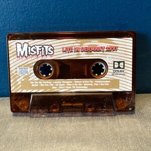 May include: A brown cassette tape with the band name "Misfits" and the text "Live in Germany 1997" printed on the front. The cassette tape has a Dolby logo and a list of song titles printed on the back.