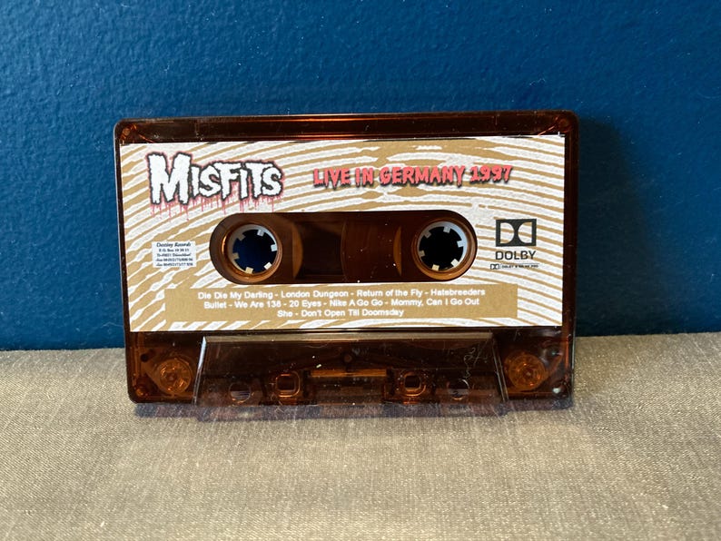 May include: A brown cassette tape with the band name "Misfits" and the text "Live in Germany 1997" printed on the front. The cassette tape has a Dolby logo and a list of song titles printed on the back.