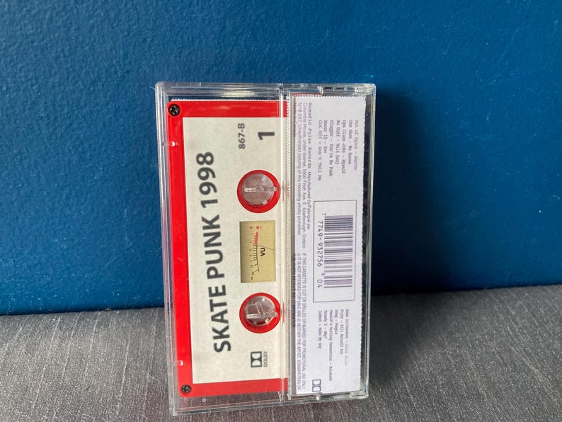 K&ouml;nnte beinhalten: Ein rotes Kassettenband mit dem Text "Skate Punk 1998" und einem Lautst&auml;rkemessger&auml;t. Das Band befindet sich in einem durchsichtigen Plastikgeh&auml;use.