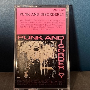 Op de afbeelding: Een vintage cassettebandje met een roze en zwarte hoes. De tekst "PUNK AND DISORDERLY" staat prominent afgebeeld, samen met een lijst van bands. De afbeelding toont een zwart-wit foto van een groep mensen.