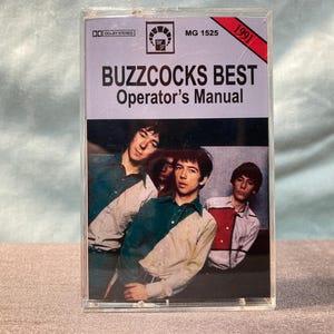 May include: A vintage cassette tape case featuring the album "Buzzcocks Best Operator's Manual" from 1991. The cover displays a band photo and the album title in bold black letters. The case is clear plastic.