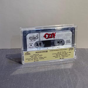K&ouml;nnte beinhalten: Eine Vintage-Audiokassette in einer transparenten Kunststoffh&uuml;lle. Das Band tr&auml;gt den Namen "Ozzy" in roter Schrift. Das Kassettenetikett und die H&uuml;lle haben zus&auml;tzlichen Text und Logos. Das Band ist ein Musikalbum.