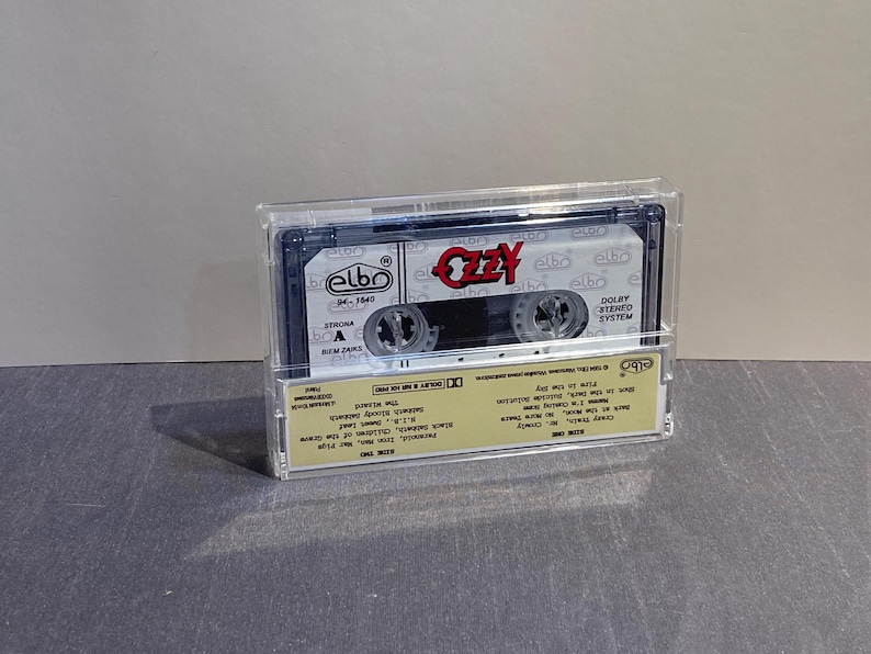 K&ouml;nnte beinhalten: Eine Vintage-Audiokassette in einer transparenten Kunststoffh&uuml;lle. Das Band tr&auml;gt den Namen "Ozzy" in roter Schrift. Das Kassettenetikett und die H&uuml;lle haben zus&auml;tzlichen Text und Logos. Das Band ist ein Musikalbum.