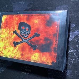 Könnte beinhalten: Eine schwarze Box mit einem Totenkopf- und Knochenkreuz-Motiv auf einem feurigen Hintergrund. Der Text "The International Heavy Metal Box Set" ist auf der Box aufgedruckt.