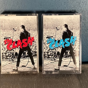 May include: Two cassette tapes with clear plastic cases. The front of each tape features a black and white photo of a person on stage performing in front of a crowd. The text "The Clash" is printed in red on one tape and blue on the other.