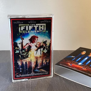 Pode incluir: Uma caixa de VHS de plástico transparente com o cartaz do filme "O Quinto Elemento". O cartaz tem um fundo vermelho e preto com imagens de atores e o título do filme. Uma caixa a condizer está ao lado.