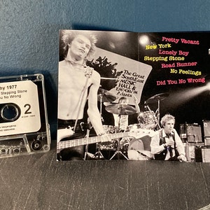 May include: A cassette tape of the Sex Pistols album "Furiously Filthy 1977". The cassette tape is in a clear plastic case. The album cover features a black and white photo of the band performing on stage. The album title is printed in white on a black background. The track list is printed in white on a black background. The album cover also includes the text "The Great South East Music Hall & Emporium Atlanta".